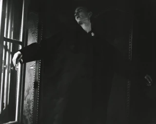 Model in a dark coat standing near a window in a dimly lit interior setting