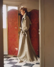 Model with spiky hair wearing a long beige belted trench coat standing before a red patterned folding screen
