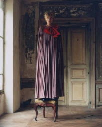 Model standing on a small wooden stool wearing a floor-length pleated purple gown with a large red bow at the neck in an ornate room.