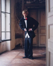 Model wearing a black suit jacket and trousers posing in an interior room with a tall window and wooden parquet floor