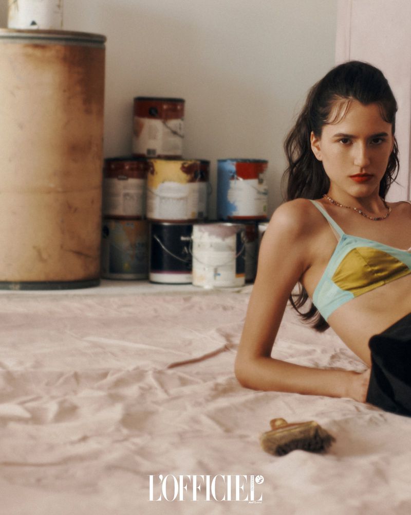 Brunette woman in a teal and gold bra lying on a pink surface in a studio with paint cans and a metal barrel in the background.