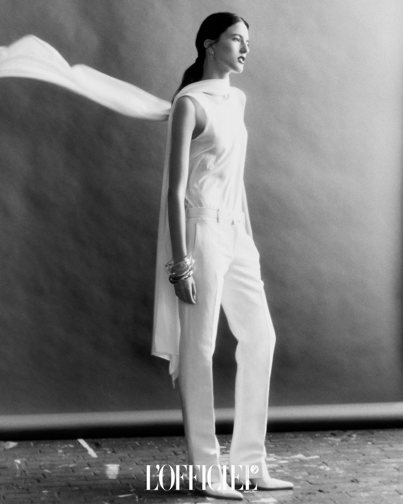 Elegant brunette woman poses in white sleeveless top and flared trousers against a mottled grey backdrop with a flowing scarf.