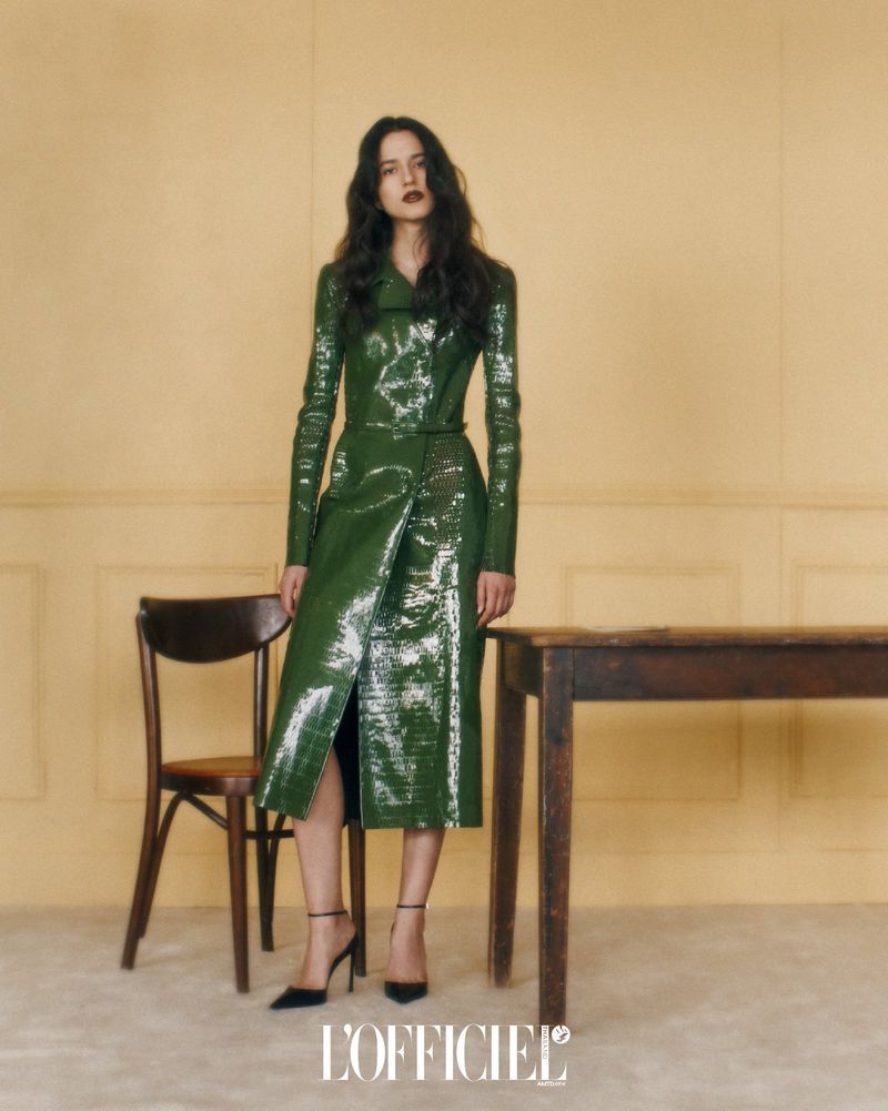 A stylish adult woman poses in a shiny green trench coat dress, black heels, and dark lipstick in a neutral-toned studio setting with chairs and a table.