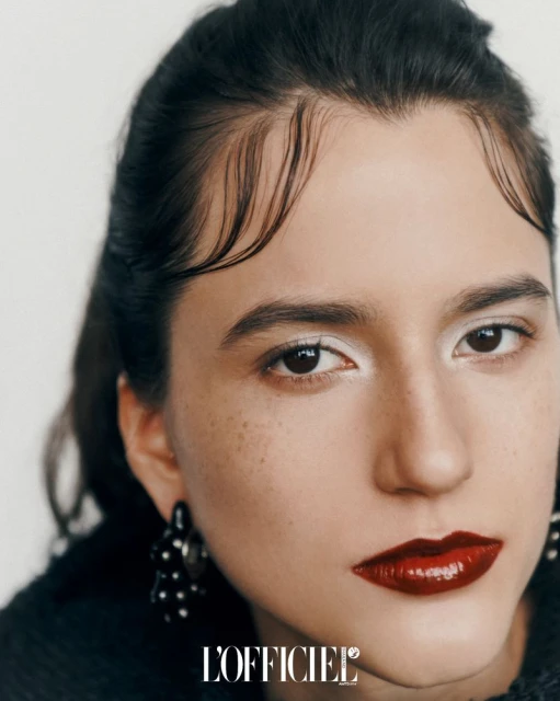 Close-up portrait of a woman with dark hair and red lipstick, posing against a white background, wearing beaded earrings and a black top.