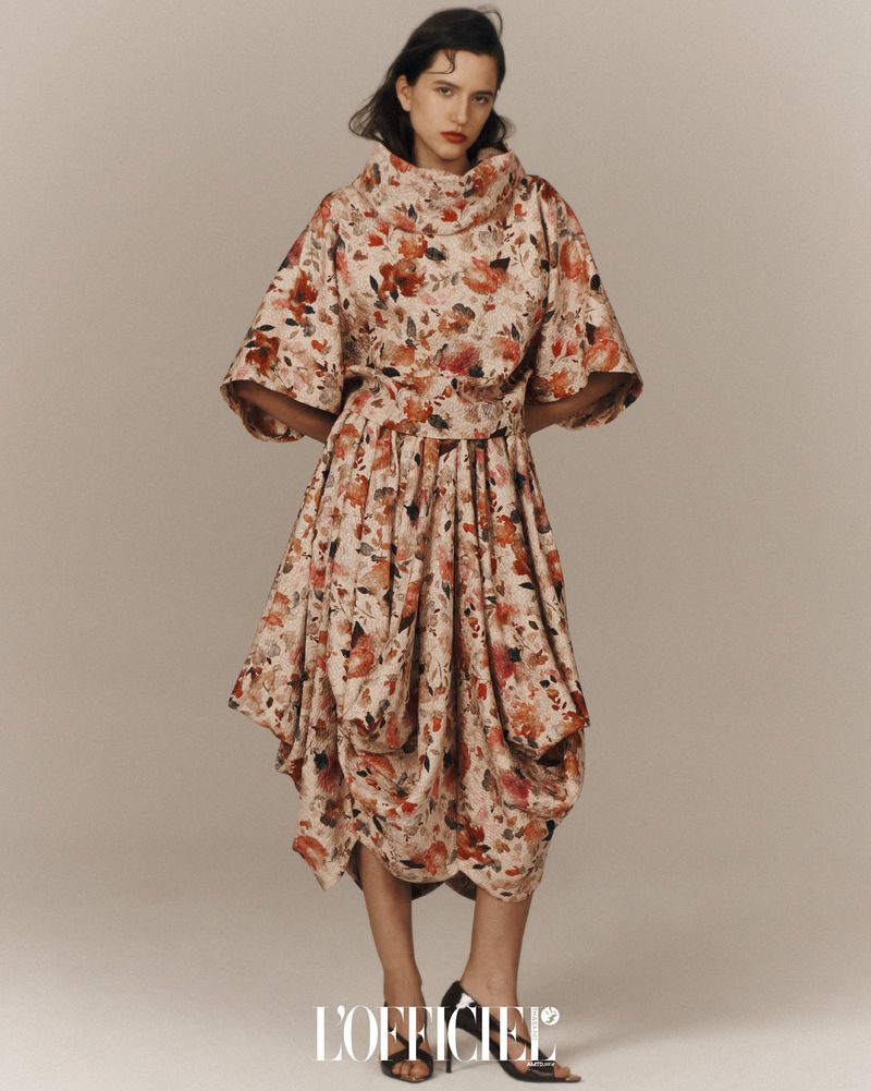 Fashion editorial shot of a woman wearing a floral print dress with a cowl neck and a cinched waist against a neutral backdrop.
