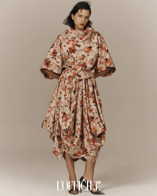 Fashion editorial shot of a woman wearing a floral print dress with a cowl neck and a cinched waist against a neutral backdrop.
