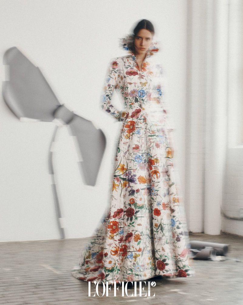 A woman poses in a full-length floral gown against a blurred background, exuding editorial fashion in an indoor studio setting.