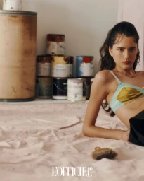 Model in a multi-colored bralette posing near stacked paint cans and a paint brush on a canvas