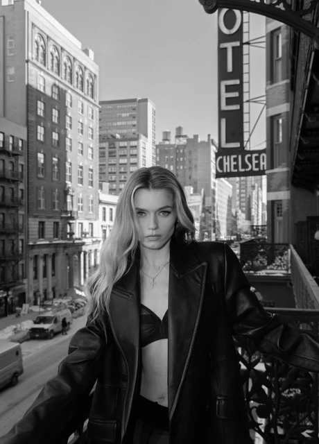 Black and white portrait of a blonde woman in a leather coat with a city street and buildings in the background.