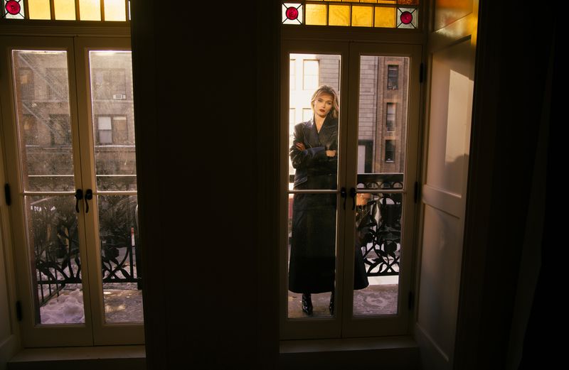 Woman in a long black coat stands on a balcony with ornate iron railings, framed by French doors in an urban setting.