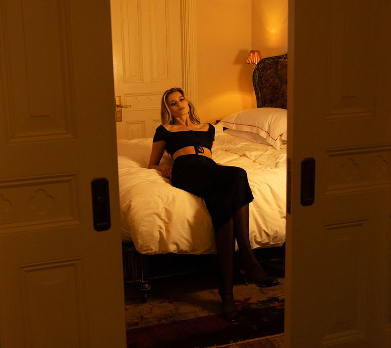 A woman in a black two-piece outfit sits on a bed in a hotel room, framed by an open doorway, with warm lighting and a cozy atmosphere.