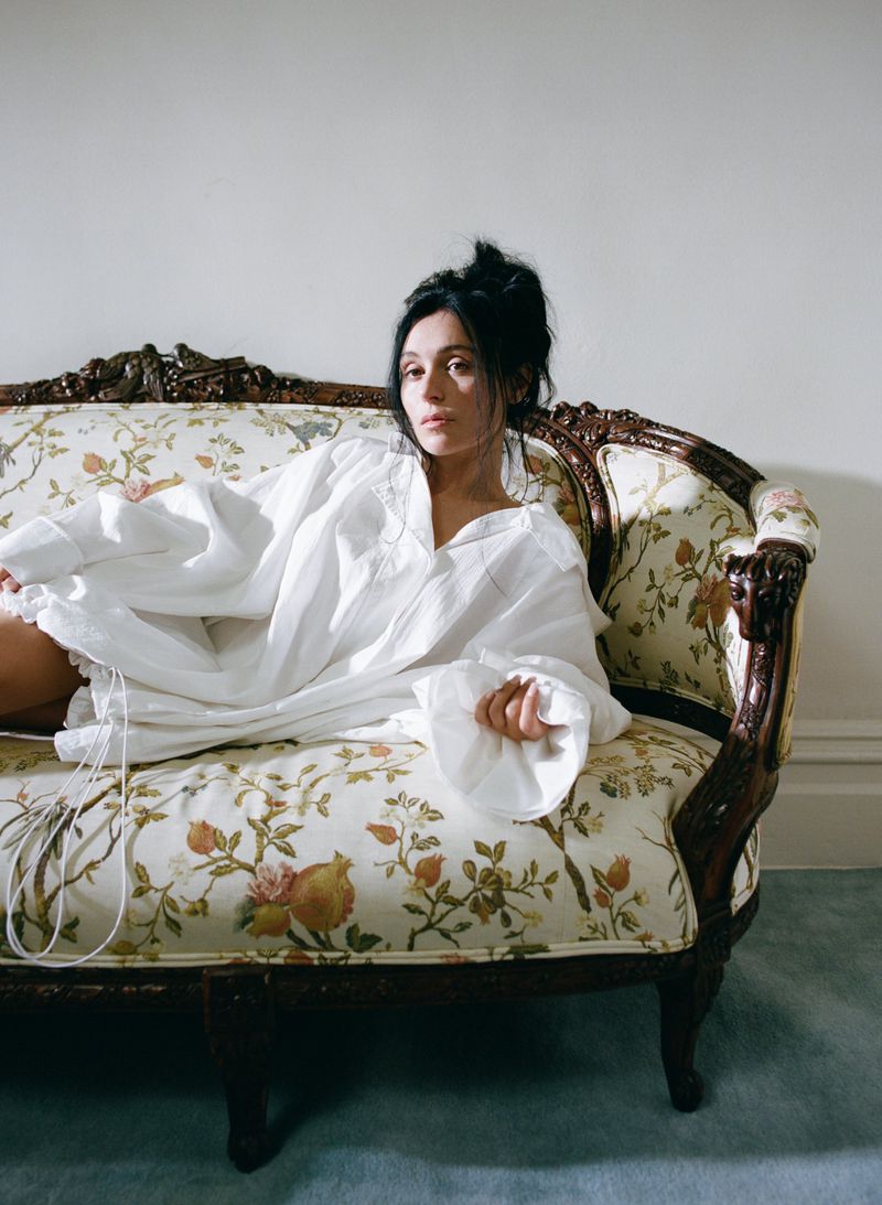 Woman reclining on a vintage floral couch in a white shirt, indoor setting. Neutral background, eye-level angle, soft diffused light.