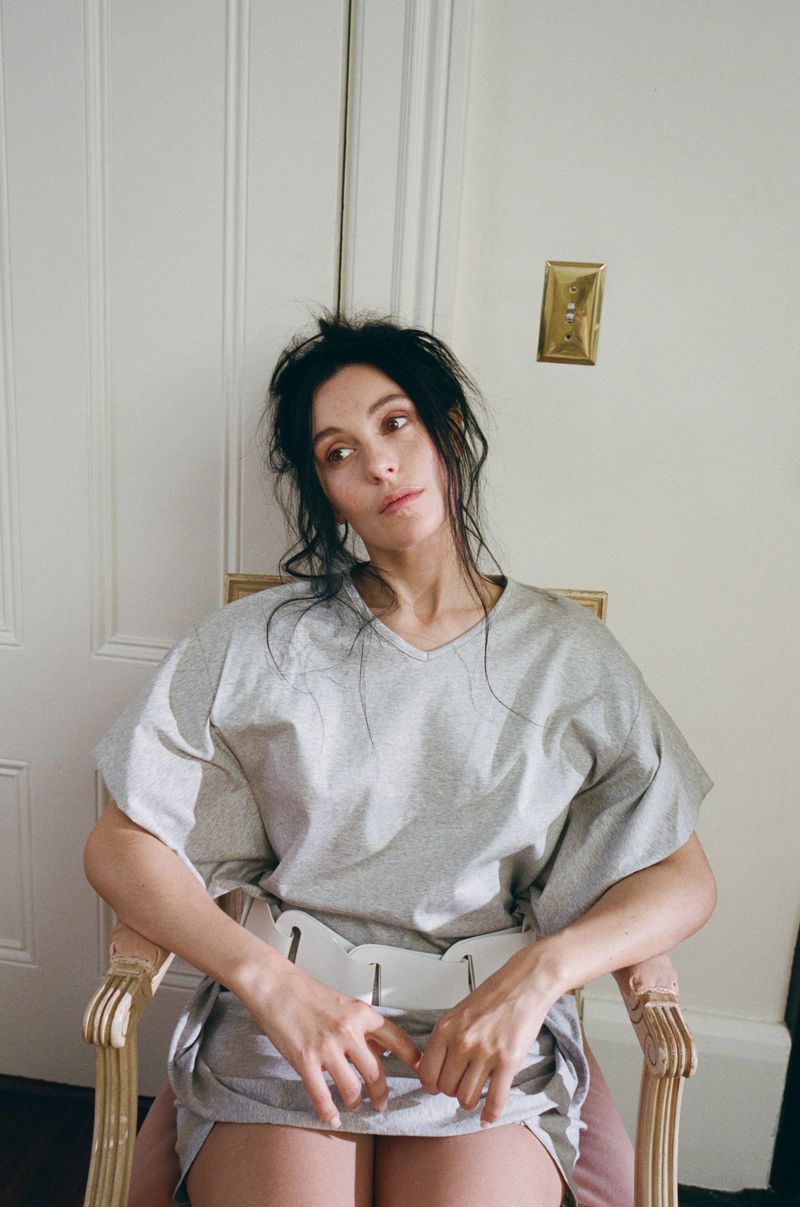 A woman sits in a chair indoors with natural light, wearing a gray top and bottom, with an off-white door and a light switch in the background.