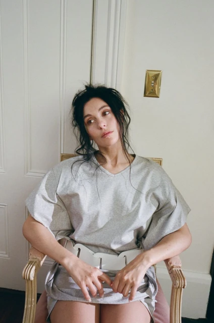 A woman sits in a chair indoors with natural light, wearing a gray top and bottom, with an off-white door and a light switch in the background.