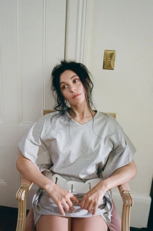 A woman sits in a chair indoors with natural light, wearing a gray top and bottom, with an off-white door and a light switch in the background.