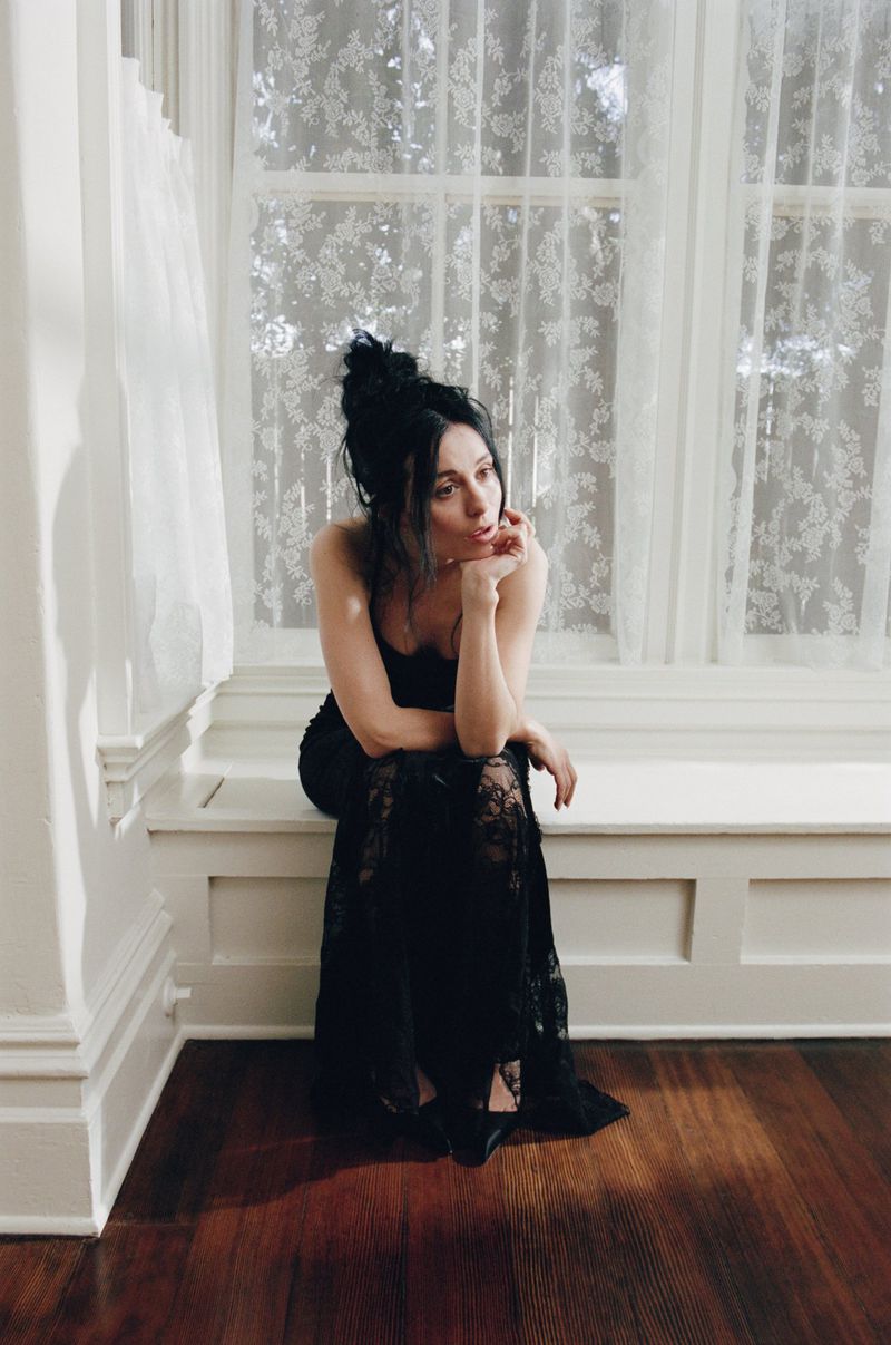 Elegant woman with black hair and a black lace dress poses while seated near a window with white lace curtains in a brightly lit room.