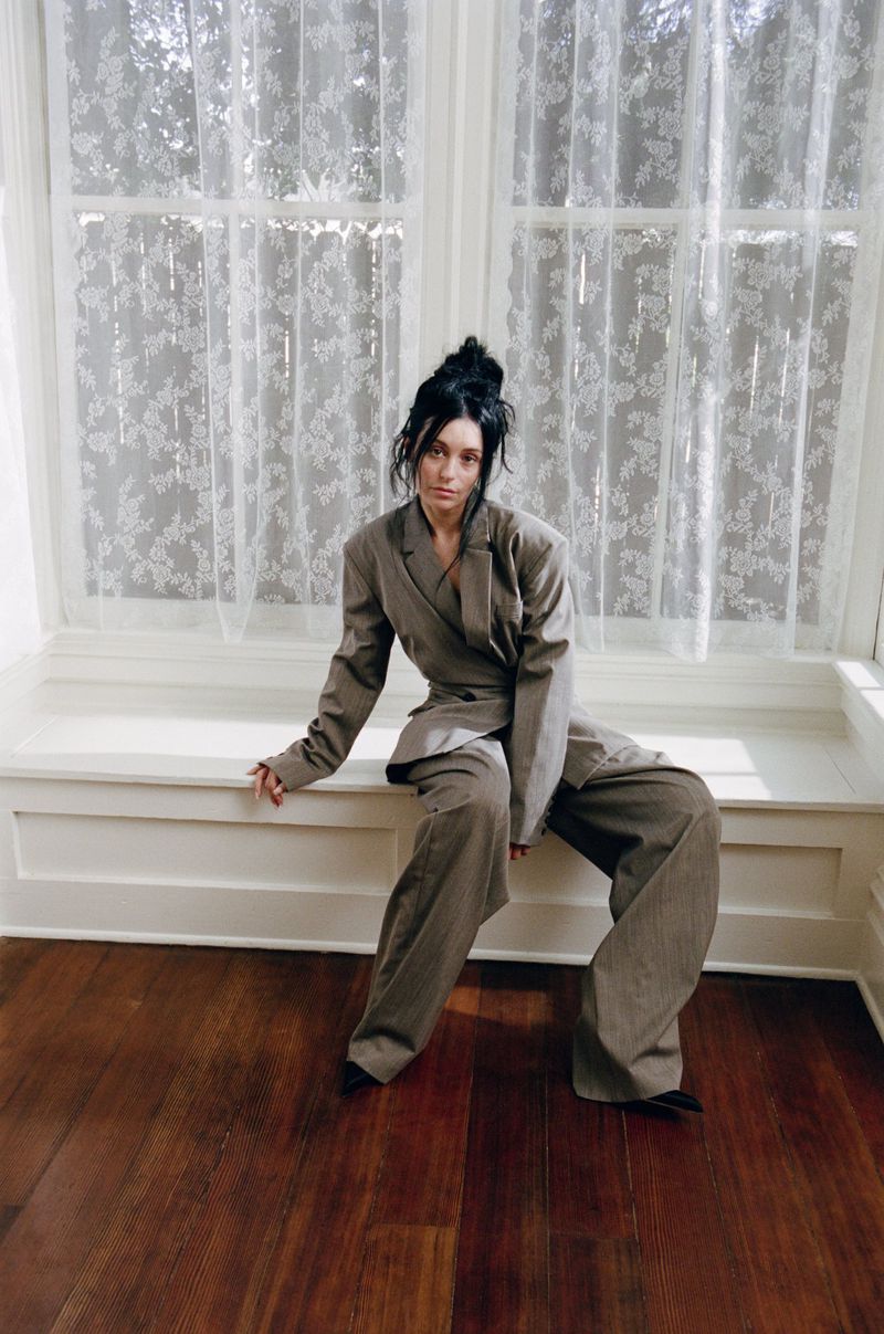 Woman in oversized grey suit sits on white window ledge in natural light with dark hair in modern fashion editorial style photo.