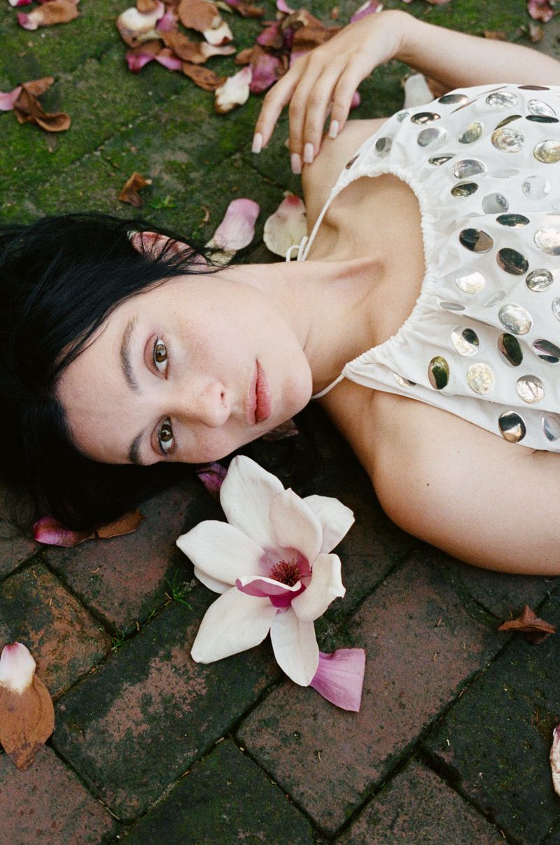 Portrait of a woman with dark hair lying on brick surface surrounded by flower petals with a large white and pink magnolia blossom.
