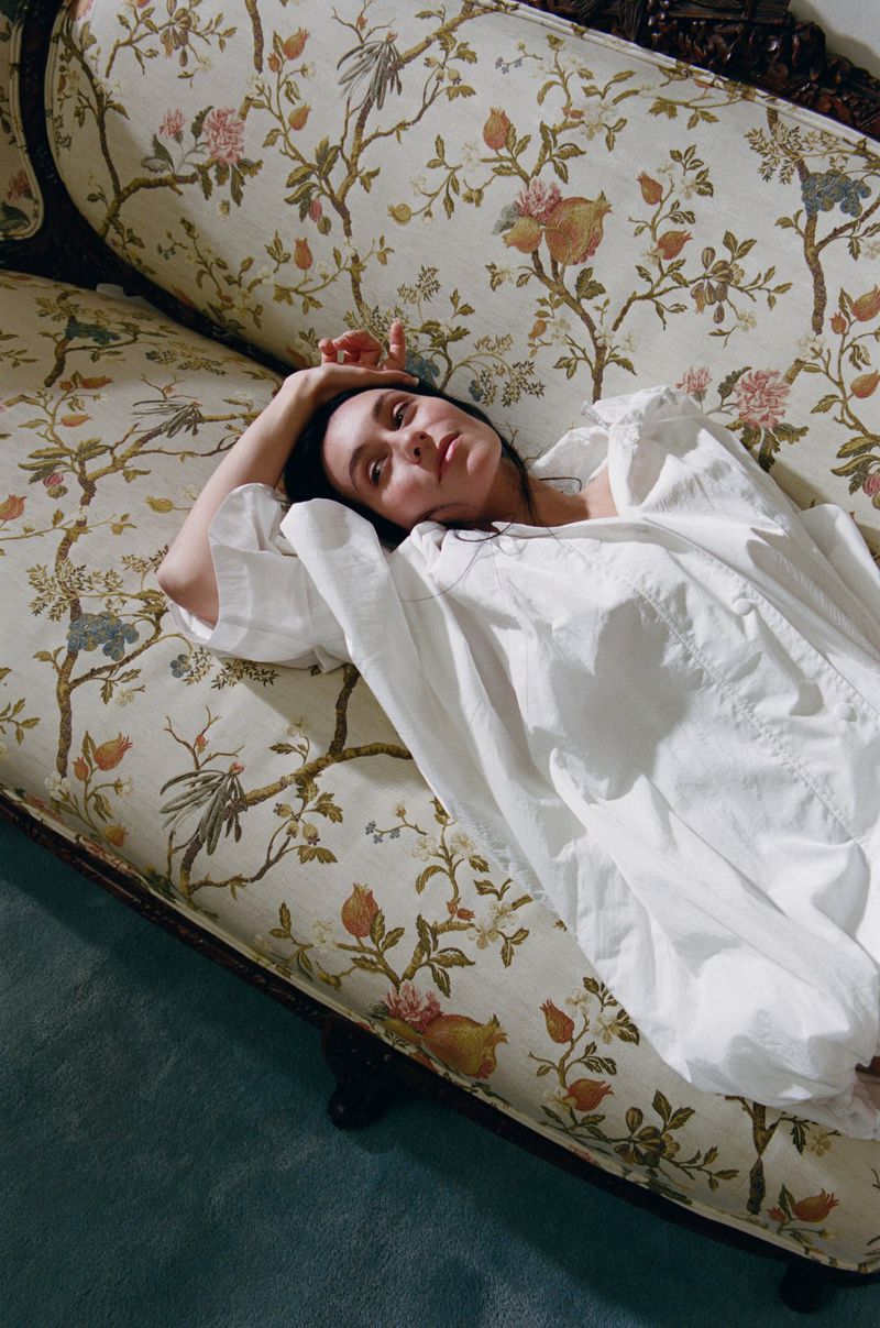 Brunette woman in white dress rests on a vintage floral couch in a calm, indoor scene featuring soft lighting and serene aesthetic.