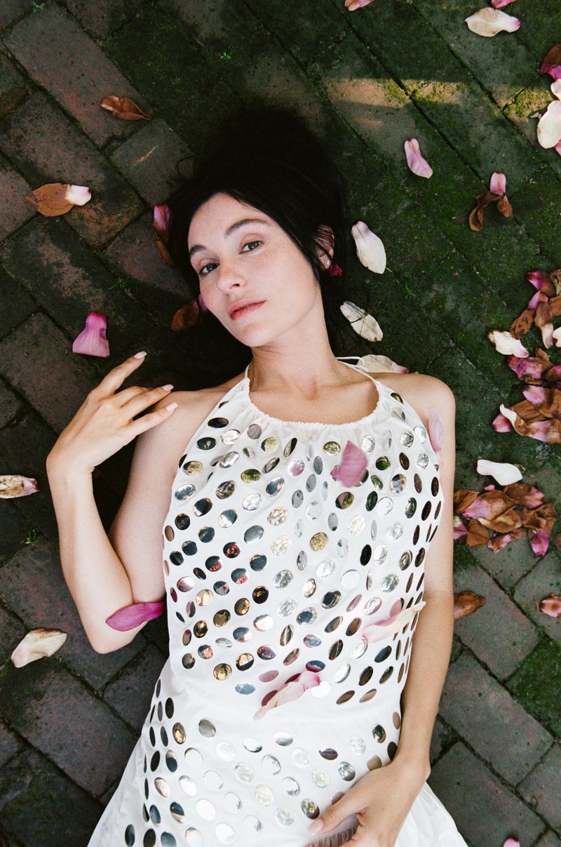 Overhead shot of a woman with dark hair lying on a brick path covered with scattered pink and white flower petals, looking at camera.