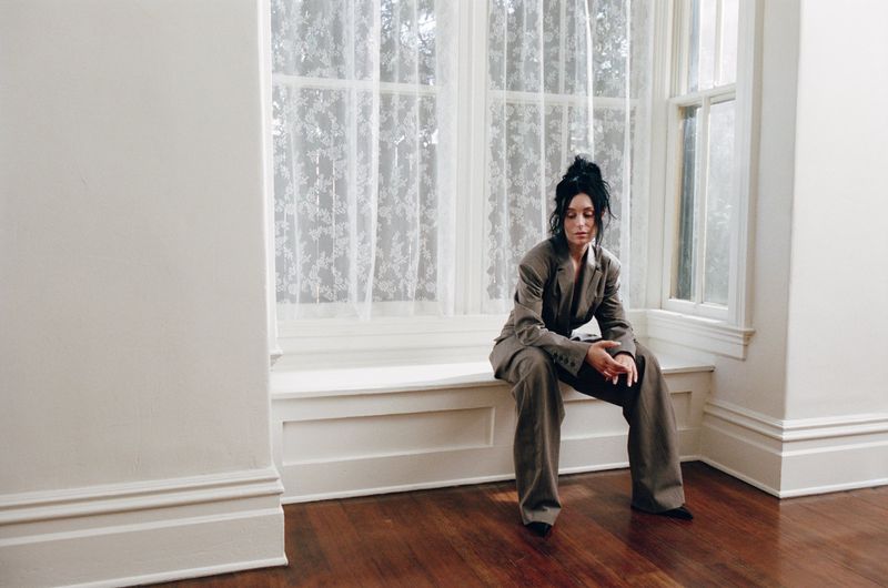 Woman in an olive green suit sitting on a window ledge in a minimalist room with hardwood floors and lace curtains in natural light.