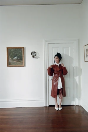 A stylish woman poses in a red leather trench coat and white heels, standing against a white door and white walls in a room with dark wood floors.