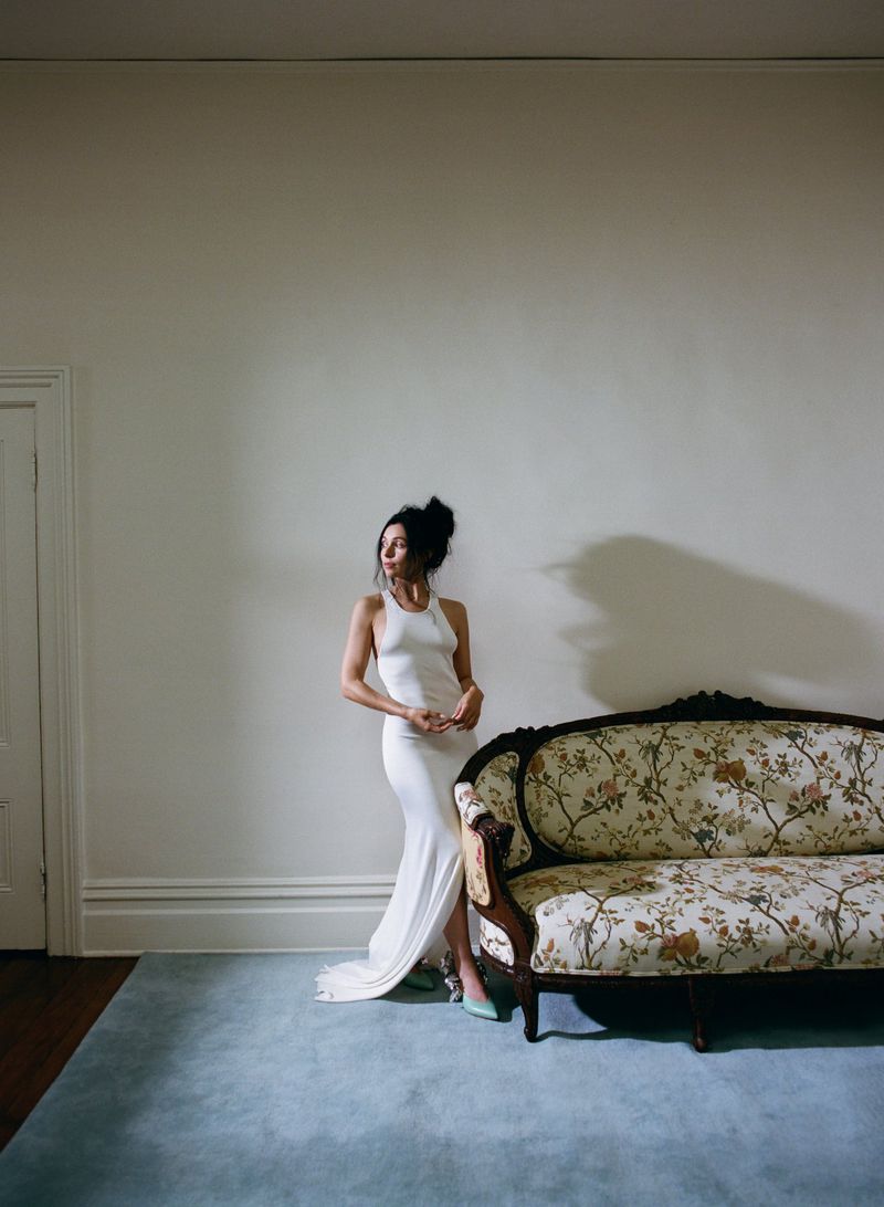 Elegant woman in white dress poses in a minimalist interior next to a vintage floral sofa, illuminated by soft natural light.