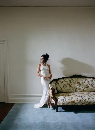 Elegant woman in white dress poses in a minimalist interior next to a vintage floral sofa, illuminated by soft natural light.