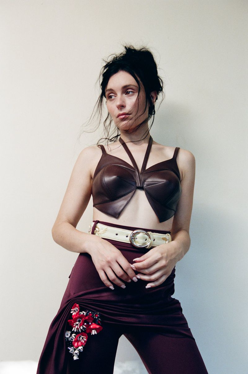 Woman in a brown leather top and maroon pants with floral detail poses against a neutral wall in a fashion portrait.