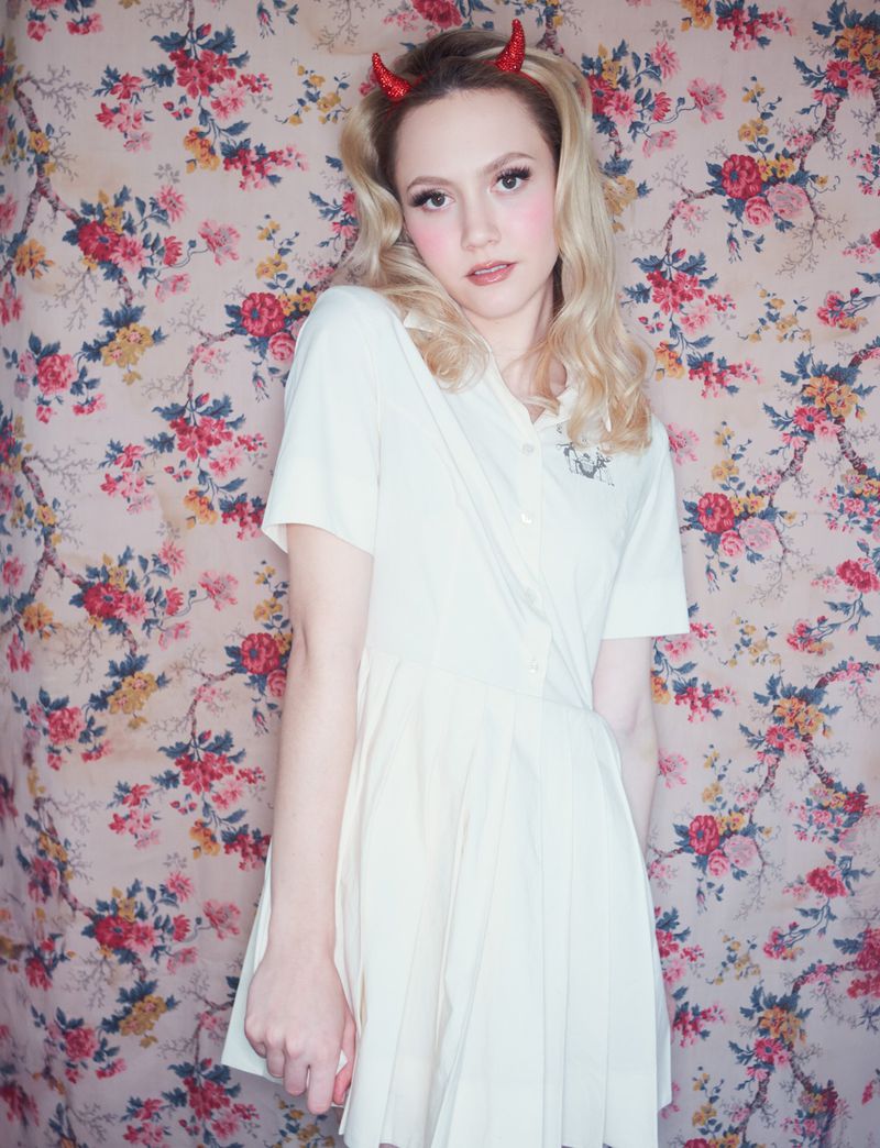 Young woman with blonde hair, pale complexion, wearing a white dress and devil horns against a floral backdrop, eye-level view.