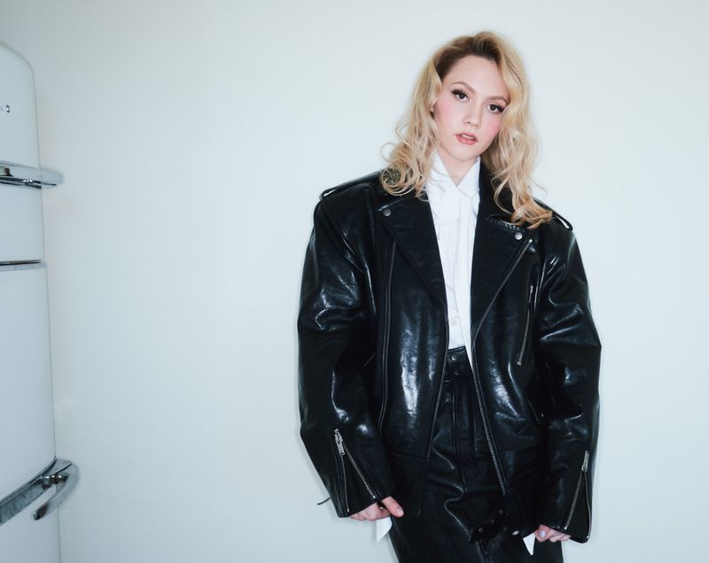 Woman in a black leather jacket and skirt poses indoors near a white refrigerator, exuding a stylish, edgy fashion aesthetic.