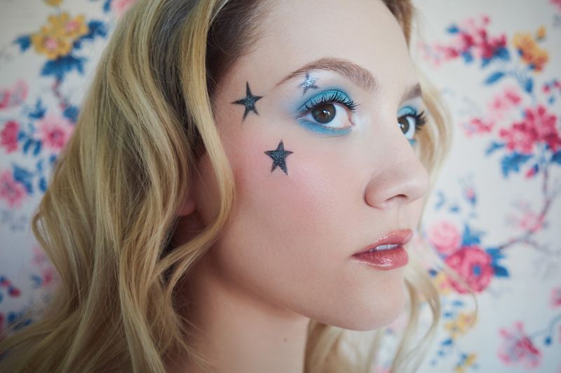 Close-up portrait of a blonde woman with blue eyeshadow and star face paint in front of a floral patterned background.