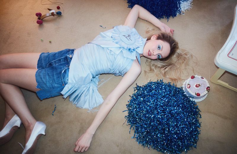 Fashion shot of a woman in a blue top and denim shorts laying on a beige floor with cake and blue tinsel around her.