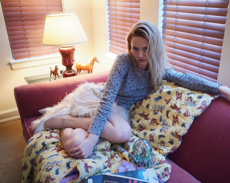 Young woman in long-sleeved shirt sits on a red couch, adorned with patterned blanket, with indoor lighting and a horse figurine.
