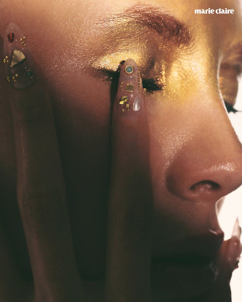 Close-up of an adult female's face with gold makeup on her eyelid and clear nail art; nail art is designed with gold shards and gems.