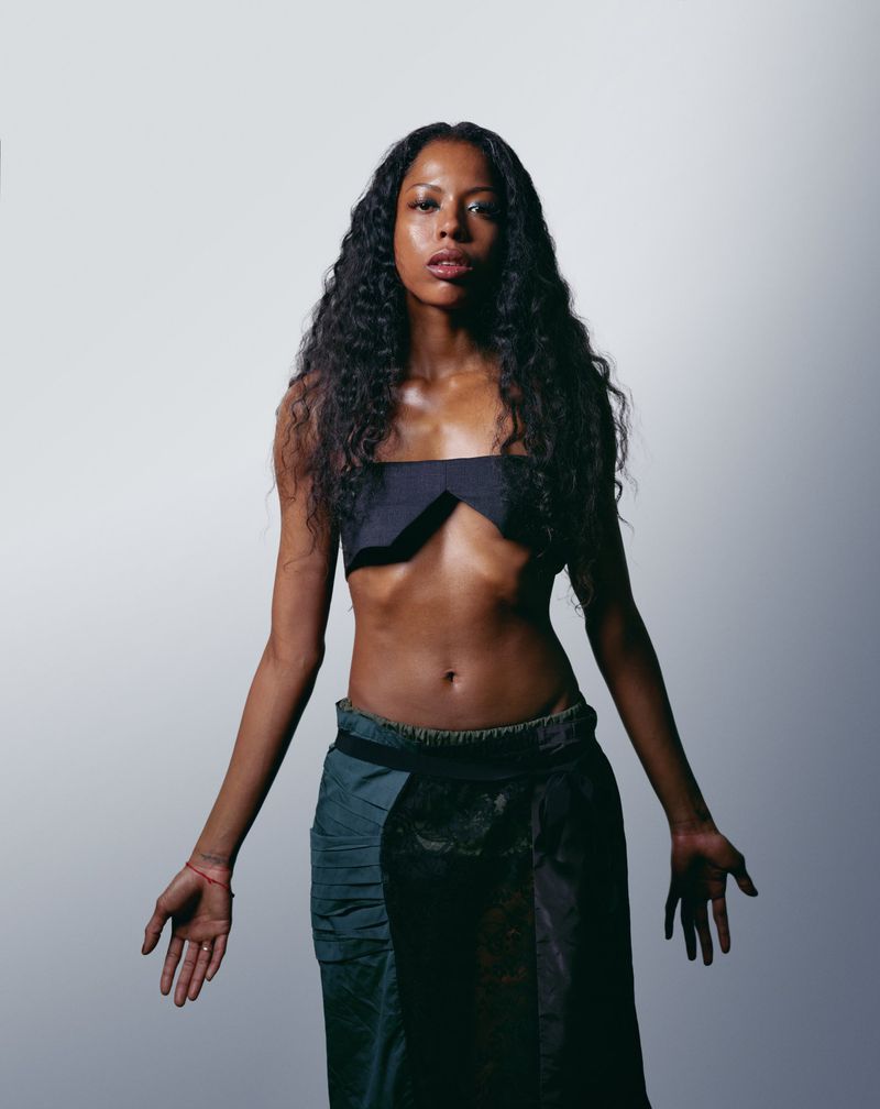 Studio portrait of a Black woman with long, curly hair wearing a black bandeau top and skirt in a front-facing pose against a white backdrop.
