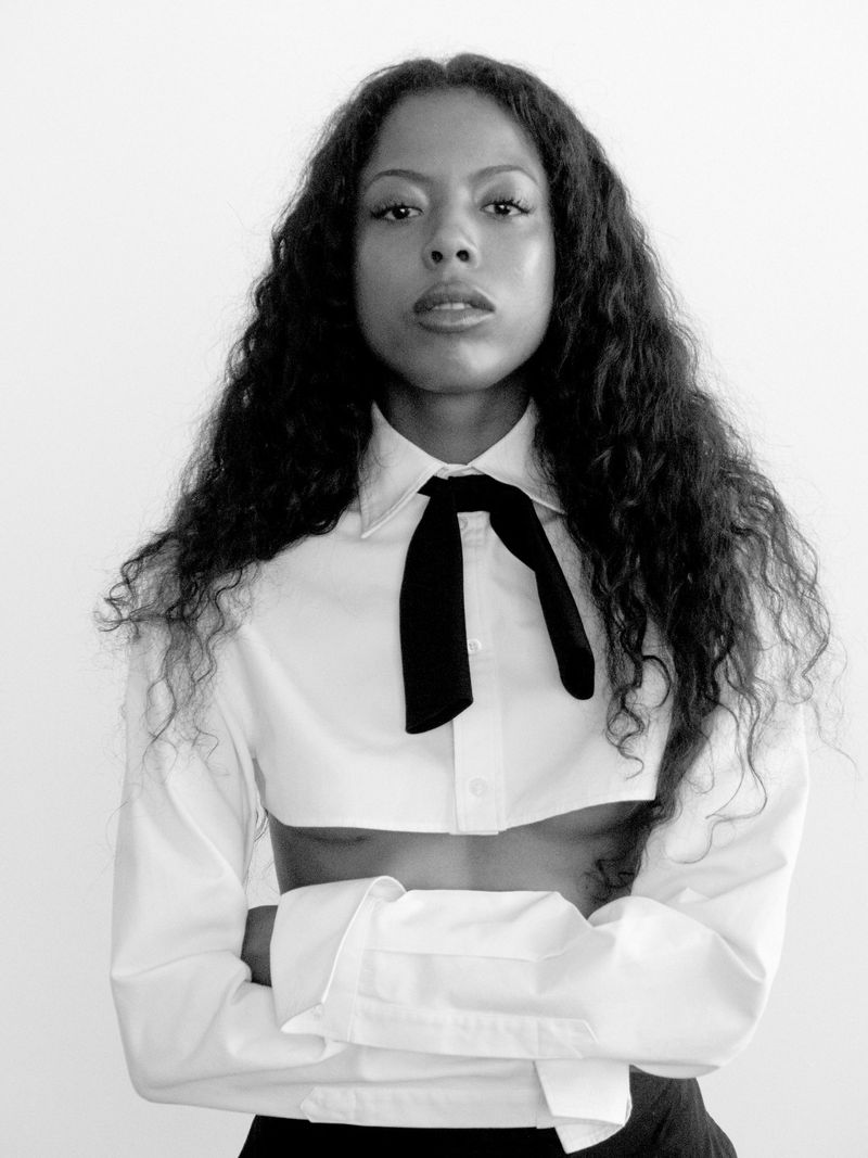 Black and white portrait of a woman with long curly hair wearing a white crop-top shirt with a black bow tie, arms crossed.