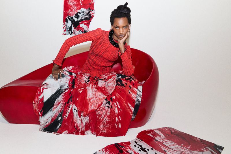 An adult woman sits in a red chair, wearing a matching red and white outfit with black accents, in a minimalistic indoor setting.