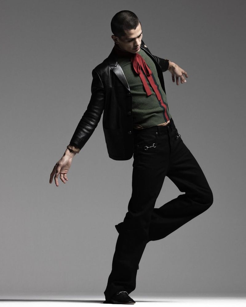 Fashionable man in black jacket and pants strikes a dynamic pose against a grey backdrop, showcasing modern style and confidence.