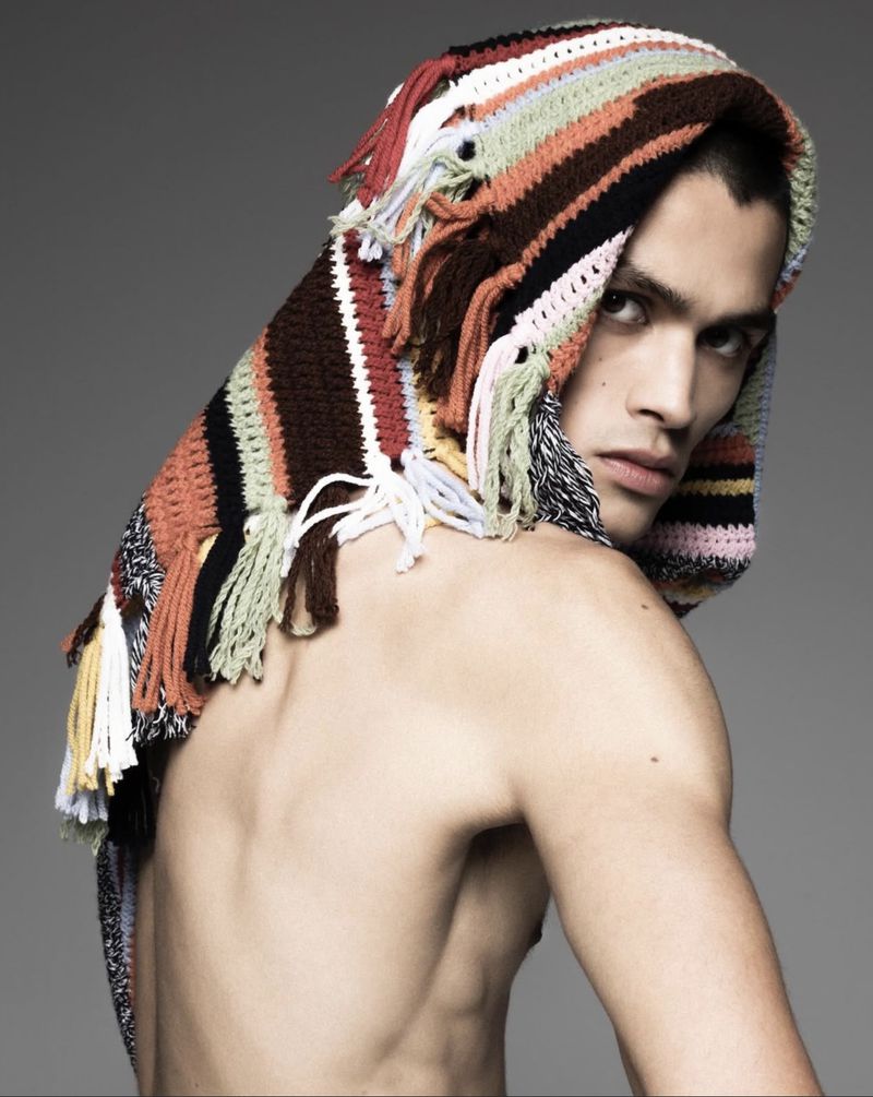 Portrait of a shirtless adult male model wearing a fringed, multicolor striped knit hood against a gray studio backdrop, close-up shot.