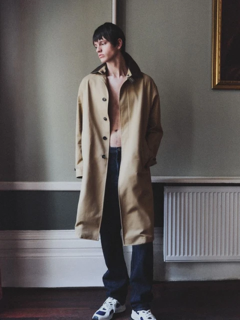 A fair-skinned male model in an unbuttoned beige trench coat, dark jeans, and sneakers poses in an old room with soft, natural lighting.
