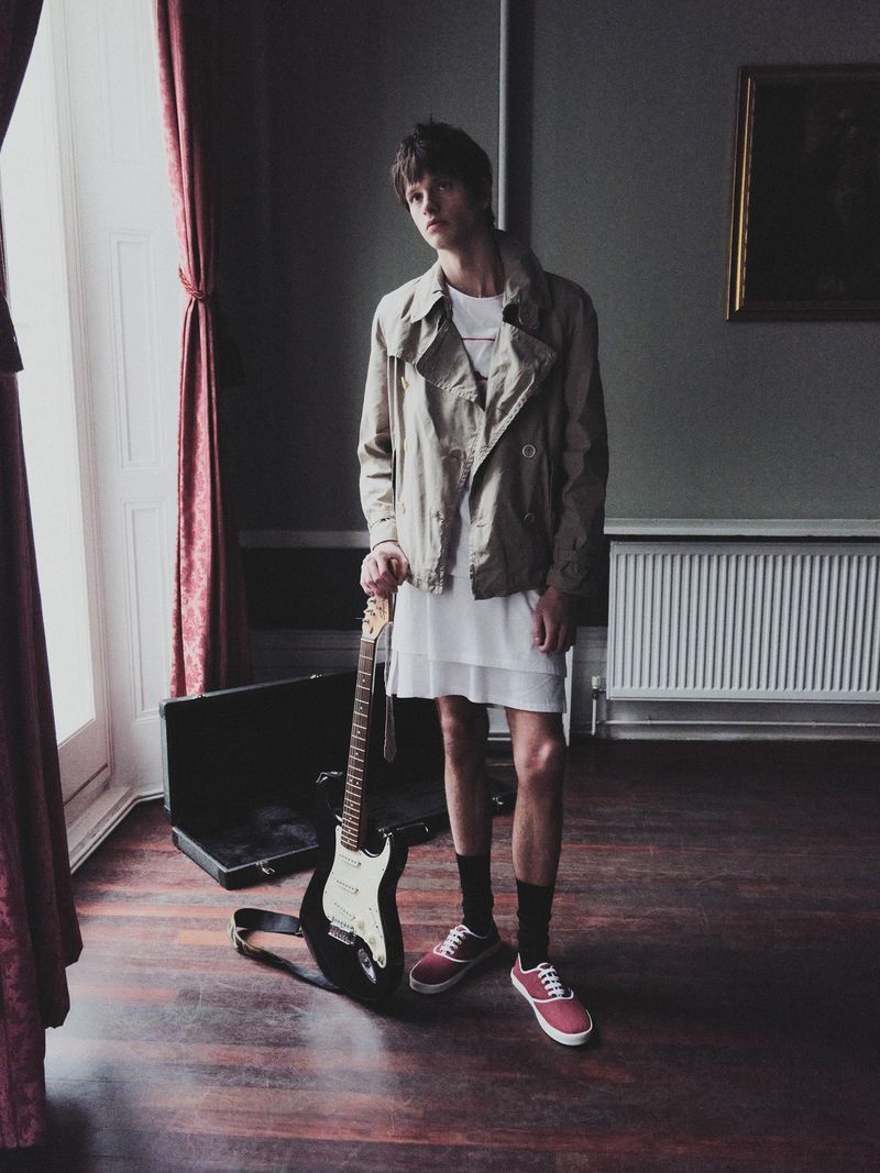 A slender young adult man with a guitar standing indoors, near a window, in a tan jacket and white dress; guitar leaning on the case.