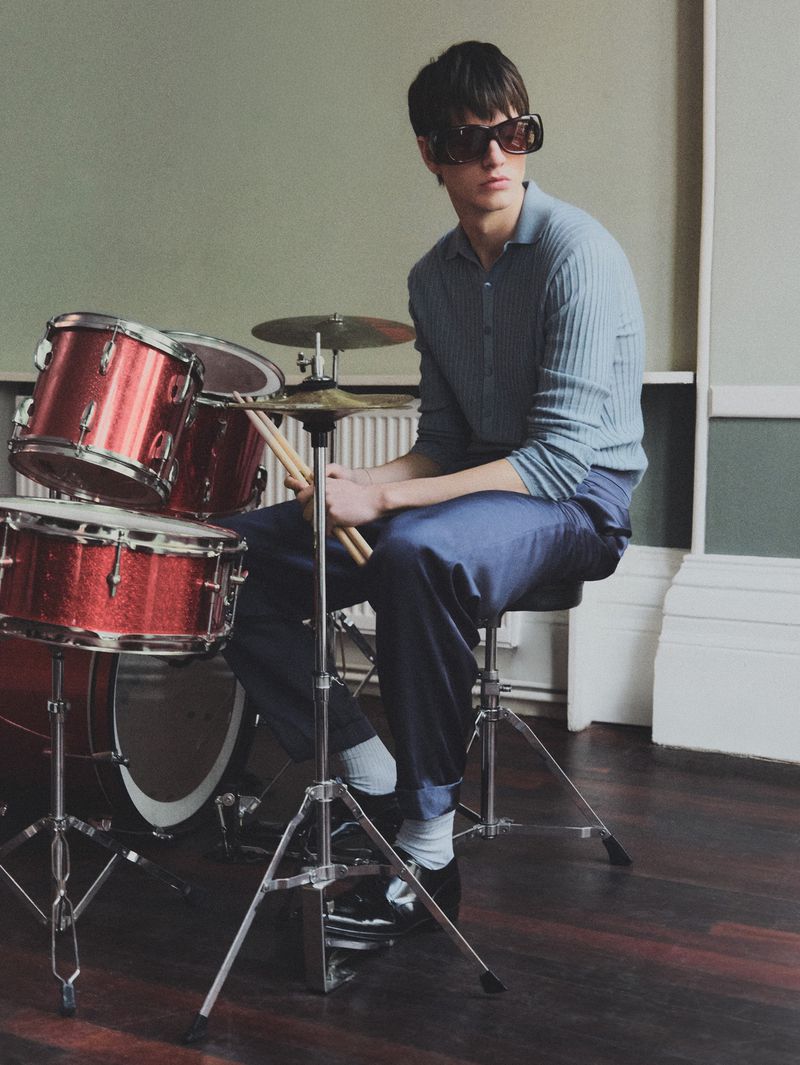A young adult male sits with a red drum set indoors, wearing a collared shirt, dark pants, and black sunglasses in a studio setting.