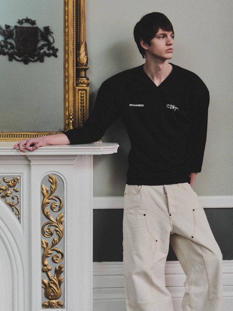 A young man poses indoors, wearing a black V-neck sweater and beige pants. He leans against a decorative fireplace with ornate gold details.