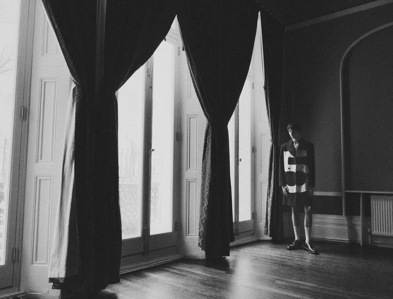 A solitary figure stands in a room with large windows draped with heavy curtains. The image is monochrome, highlighting the architectural details.