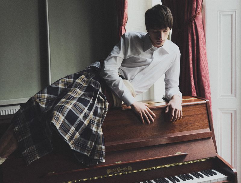 Young adult in stylish plaid kilt and white shirt sits on a wood piano in a room with window and curtains. Indoor fashion.