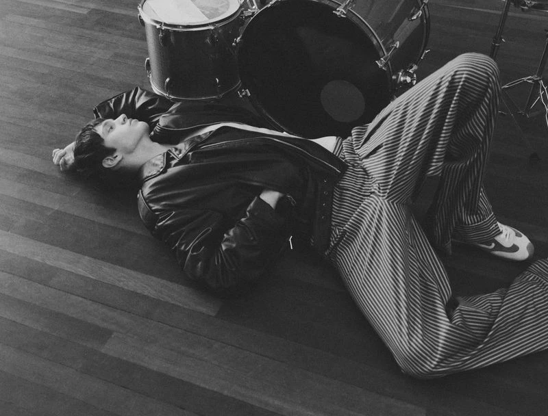 Monochrome image of a person in a leather jacket and striped pants lying on a wooden floor, with a drum kit visible in the background.