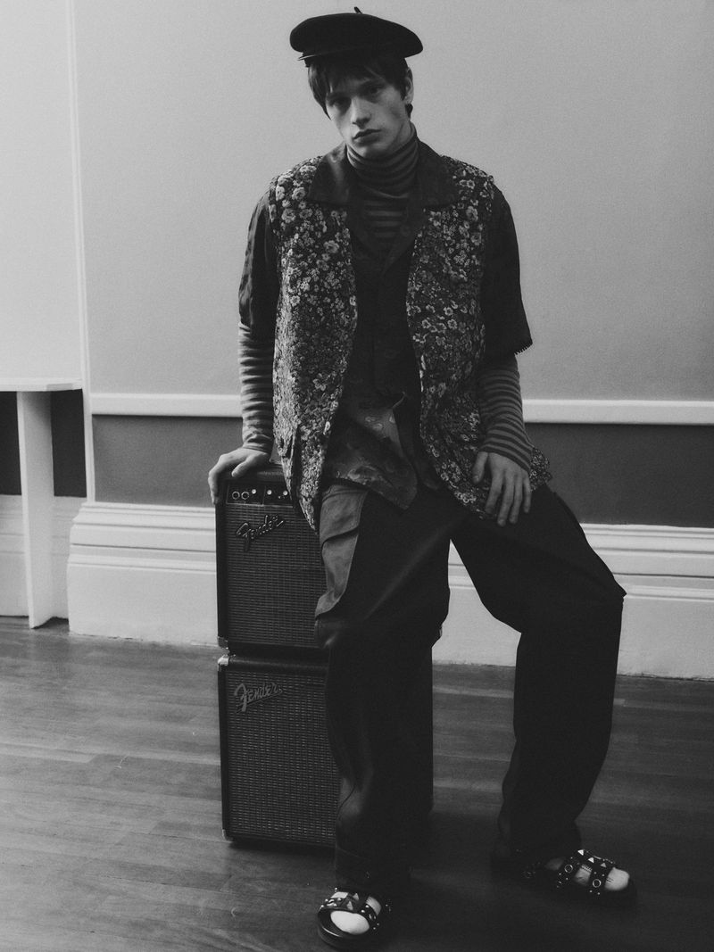 Monochrome portrait of a young adult male wearing a beret, striped shirt, and floral vest leaning against stacked Fender amps in-studio.