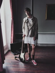 Young man holding an electric guitar standing in a room with dark wood floors and grey walls for HERO Magazine