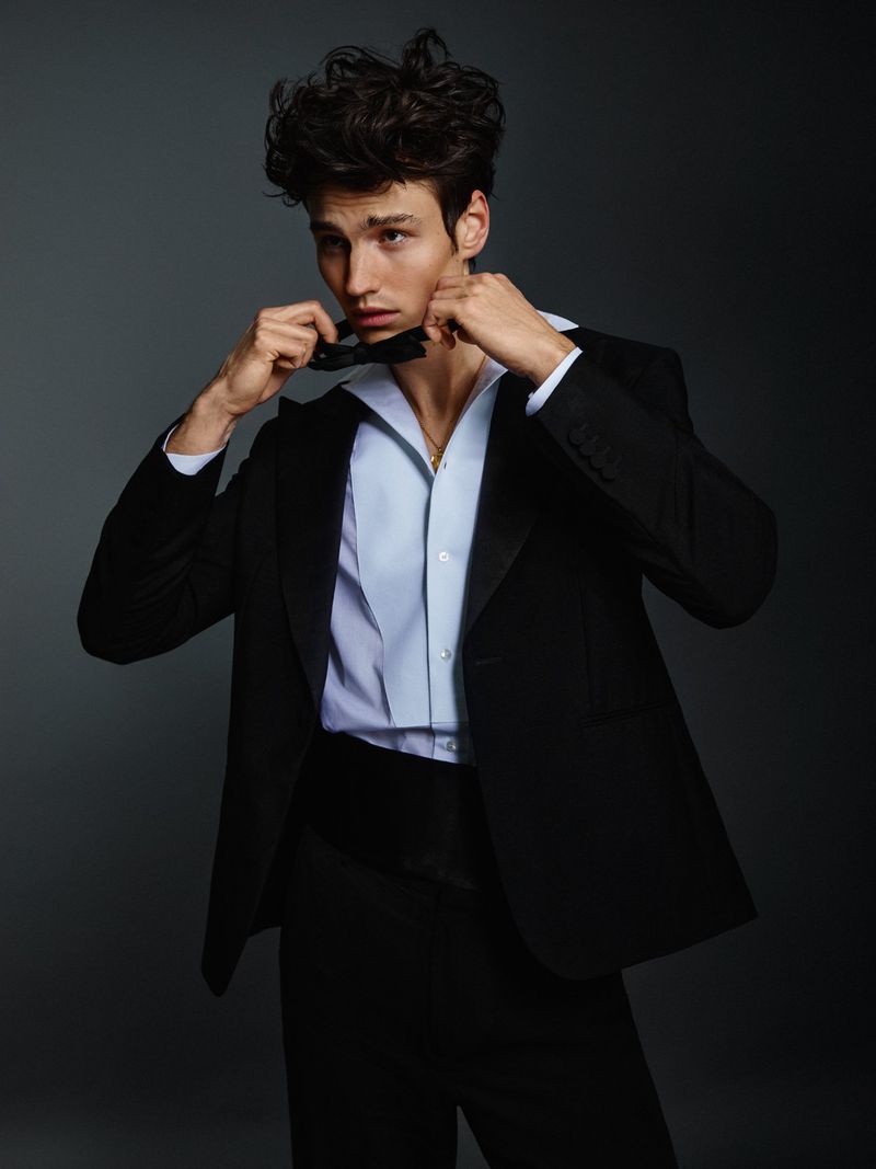 Elegant adult male poses with a black suit and bowtie against a grey background in a studio setting, showcasing formal wear.